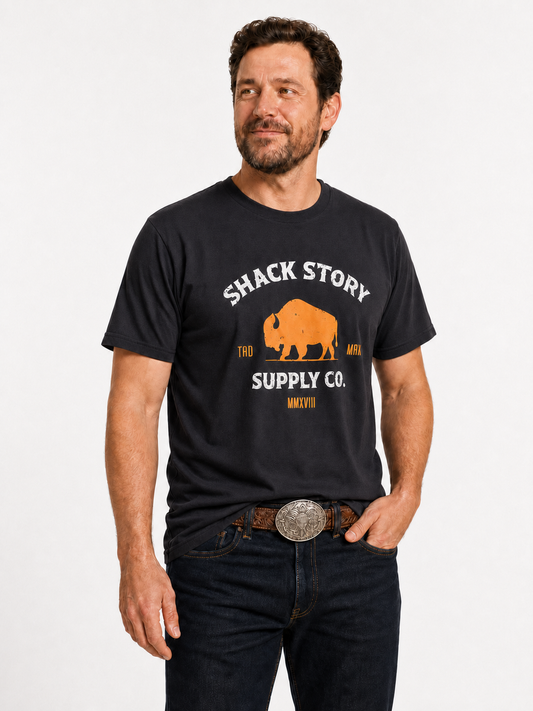Man wearing the black Shack Story Buffalo unisex T-shirt with western jeans and belt buckle, front view